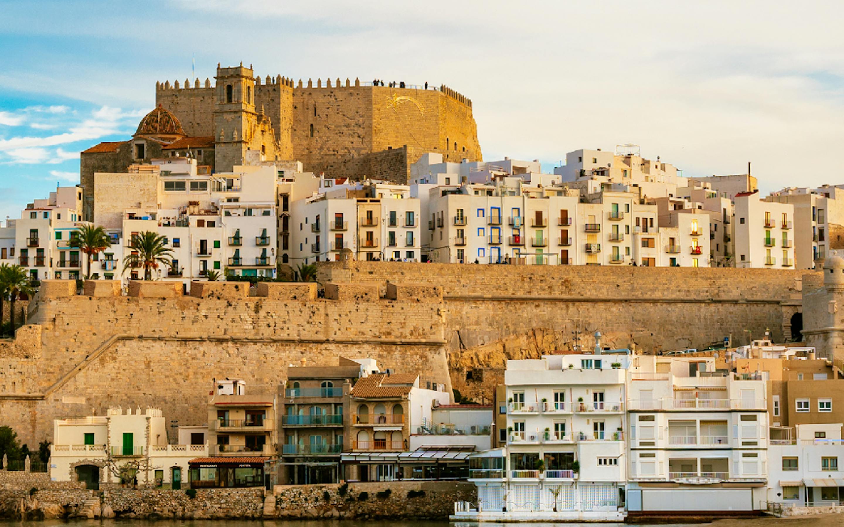 an image of an old city along the Iberian coast with beige buildings rising from the sea and a blue sky