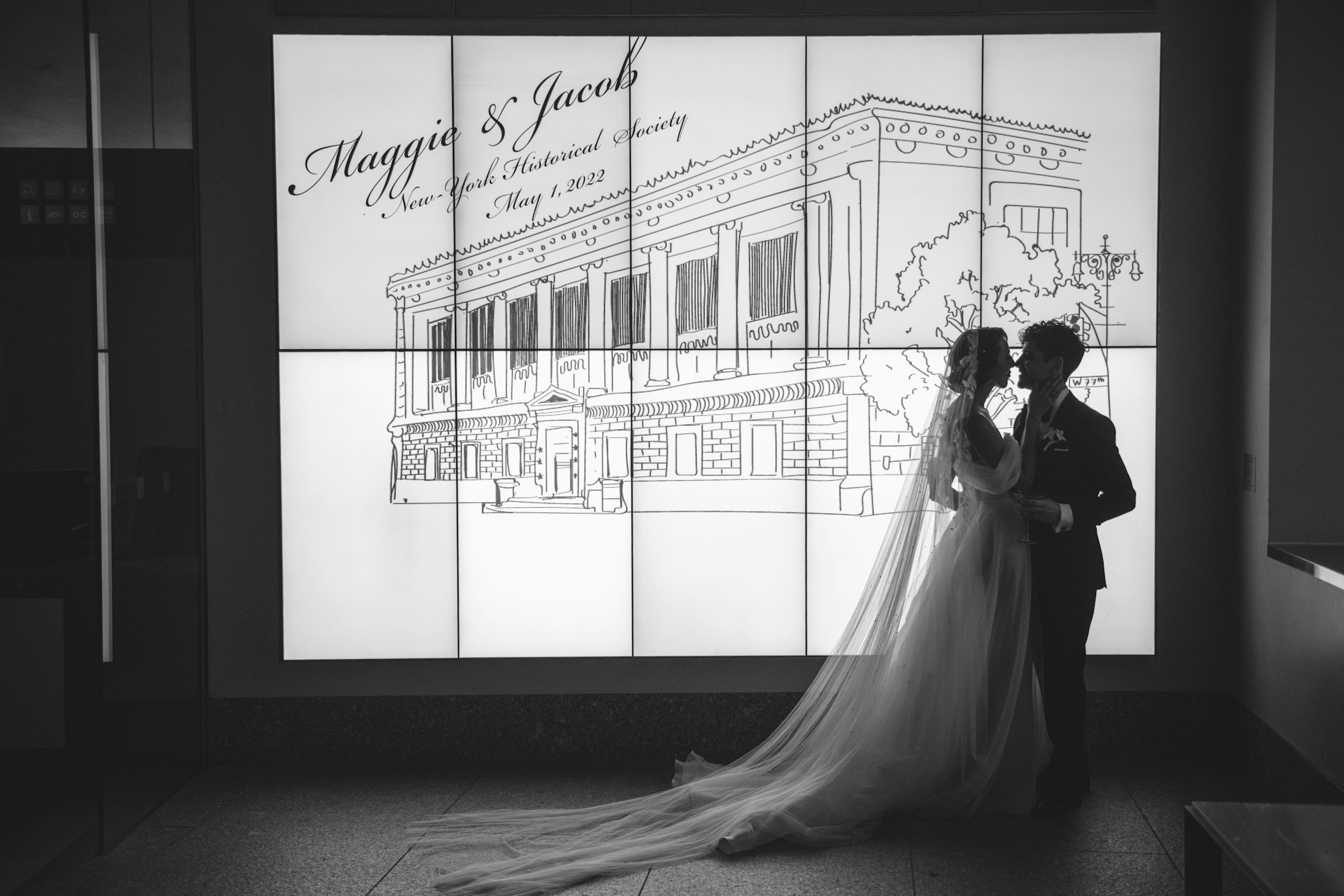a black and white image of a couple in the museum lobby in front of the digital wall showing the logo for their wedding