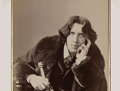 Photograph shows Oscar Wilde, three-quarter length portrait, facing front, seated, leaning forward, left elbow resting on knee, hand to chin, holding walking stick in right hand, wearing coat.