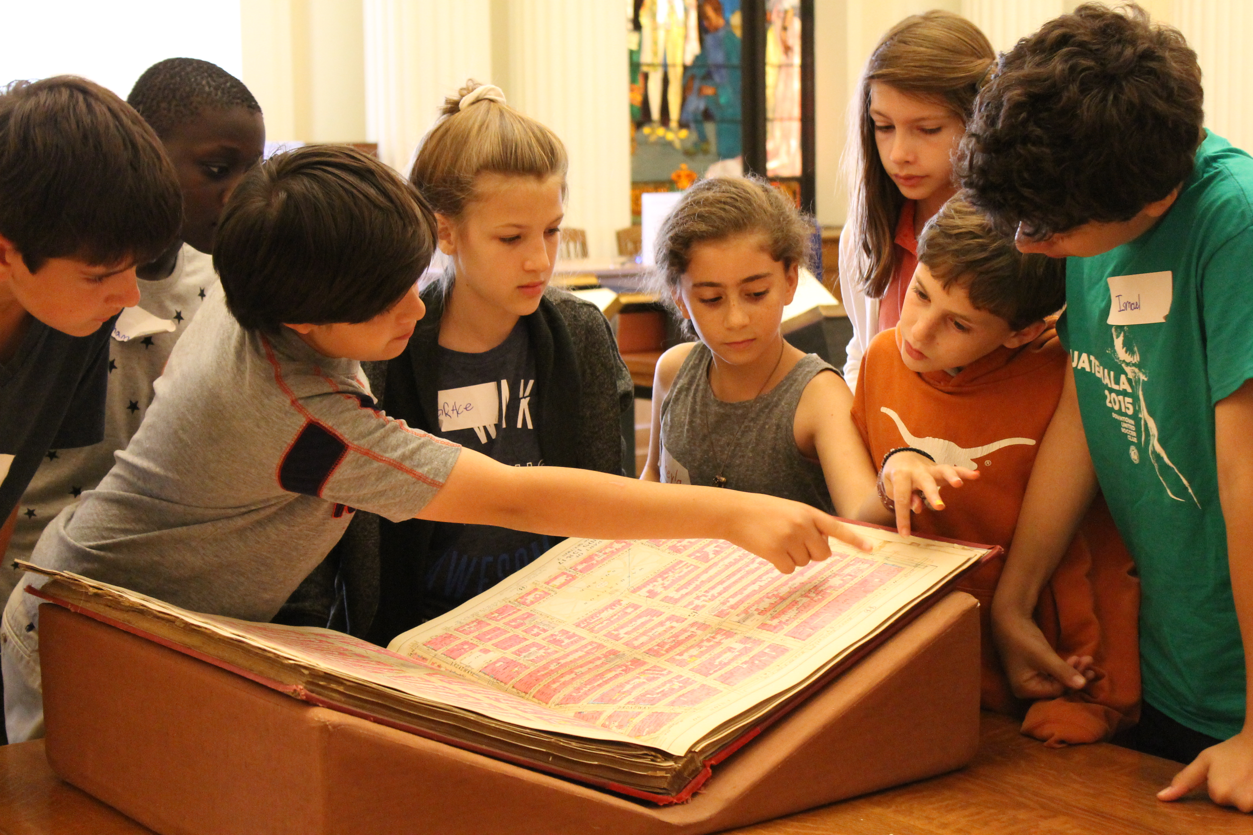 a group of kids in the museum and library look at a historic book and point at one of the pages