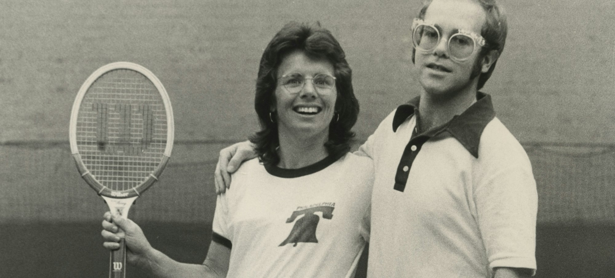 billie jean king and Elton John on a tennis court in 1974—Billie Jean is holding up a tennis racquet and they have their arms around each other.
Billie Jean King and Elton John, 1974. Courtesy of Terry O’Neill / Iconic Images Gallery