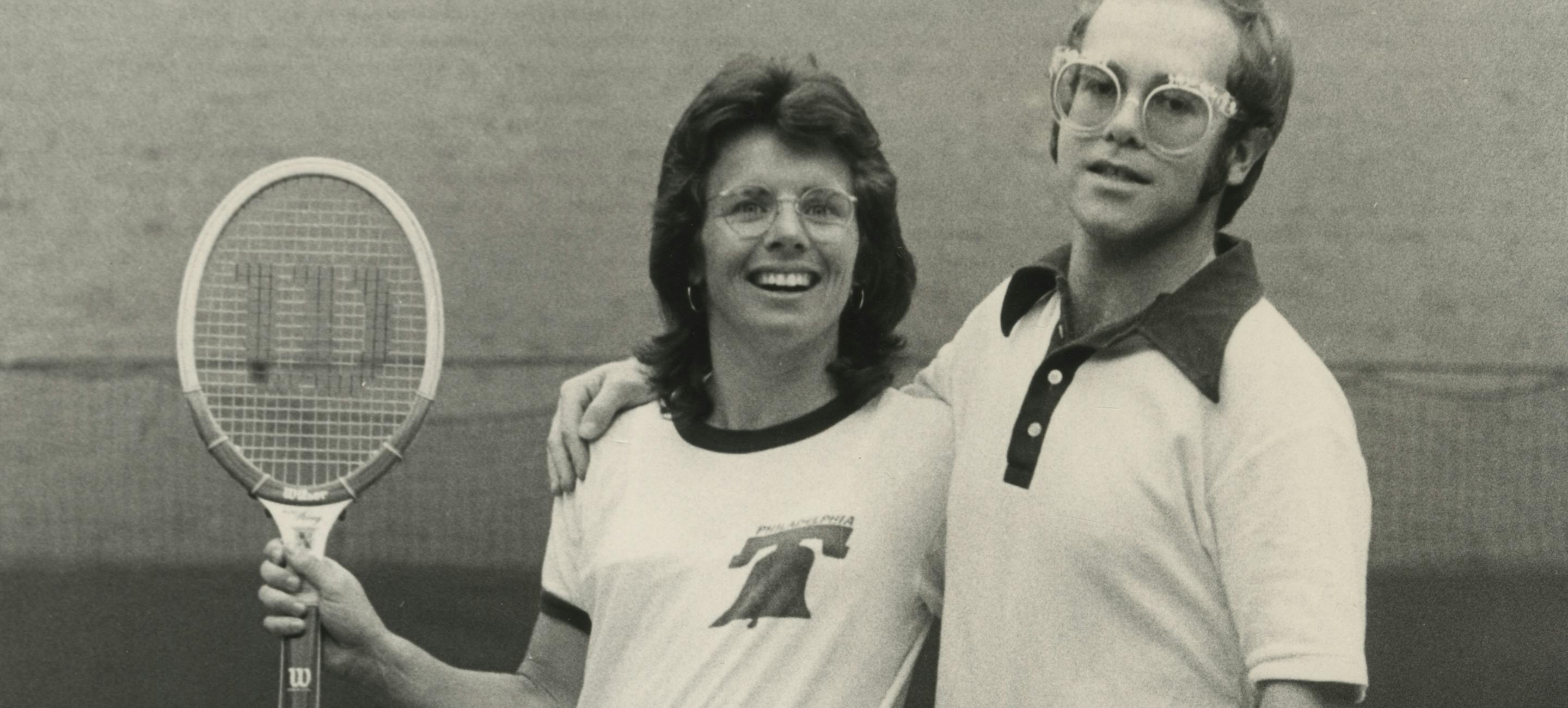 billie jean king and Elton John on a tennis court in 1974—Billie Jean is holding up a tennis racquet and they have their arms around each other.
Billie Jean King and Elton John, 1974. Courtesy of Terry O’Neill / Iconic Images Gallery