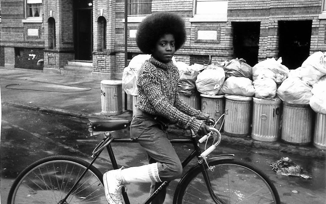 A black and white image of a boy with an afro riding a bike down a city street with full metal trash cans in the background