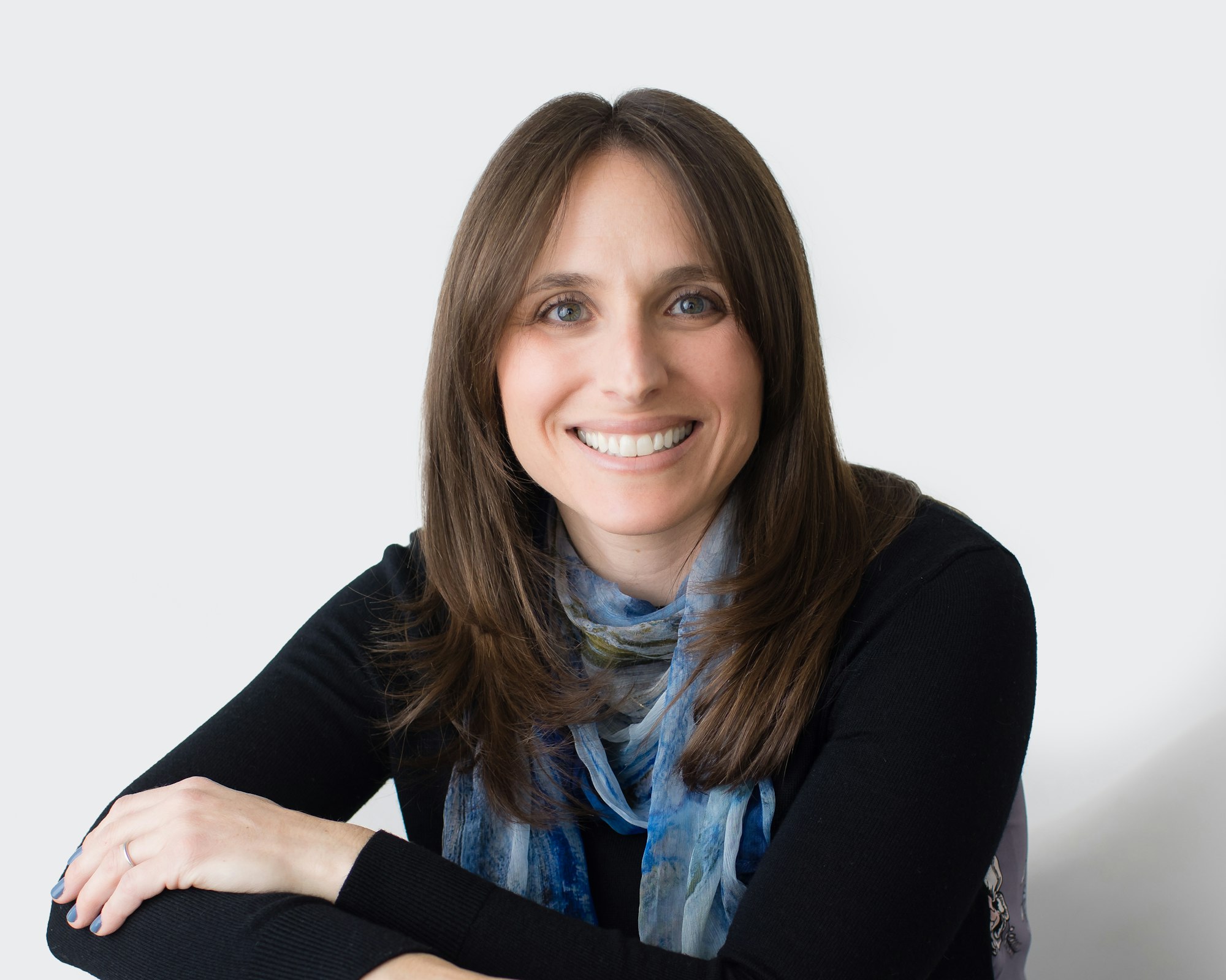 Photo of author Ami Polonsky. In this photo we see a white woman with long straight brown hair wearing a black shirt and blue scarf. She is looking and smiling at the camera.