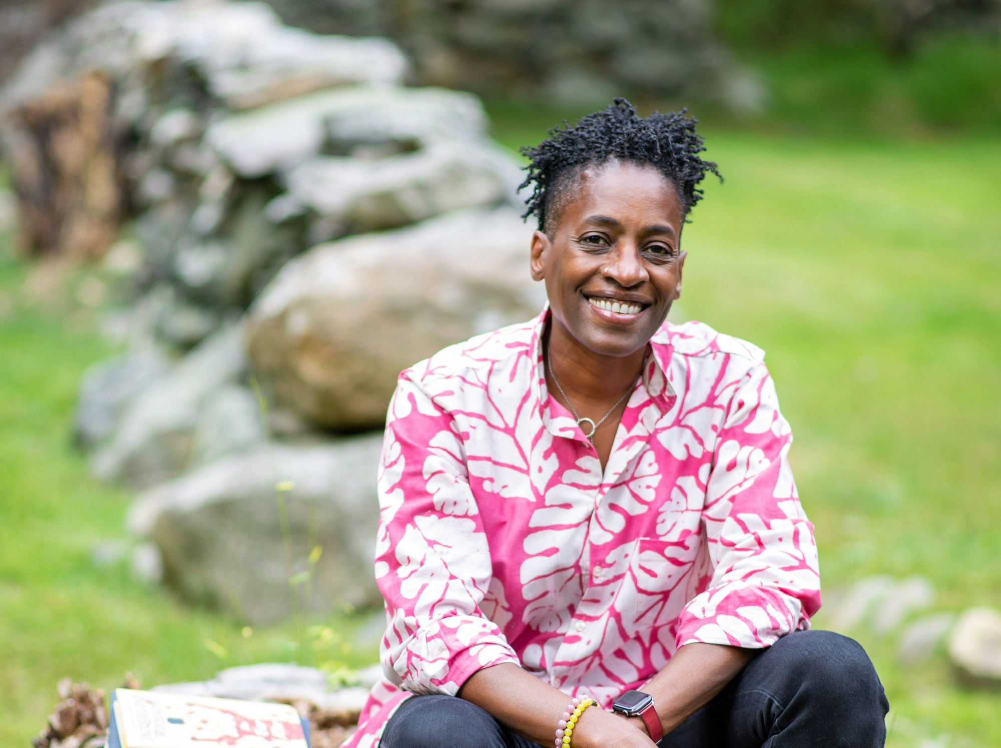 Photo of author Jacqueline Woodson. Jacqueline is a black woman with short black hair, and she is wearing a pink and white shirt with jeans. She is seated on a stump surrounded by green grass. She is smiling and facing the camera.