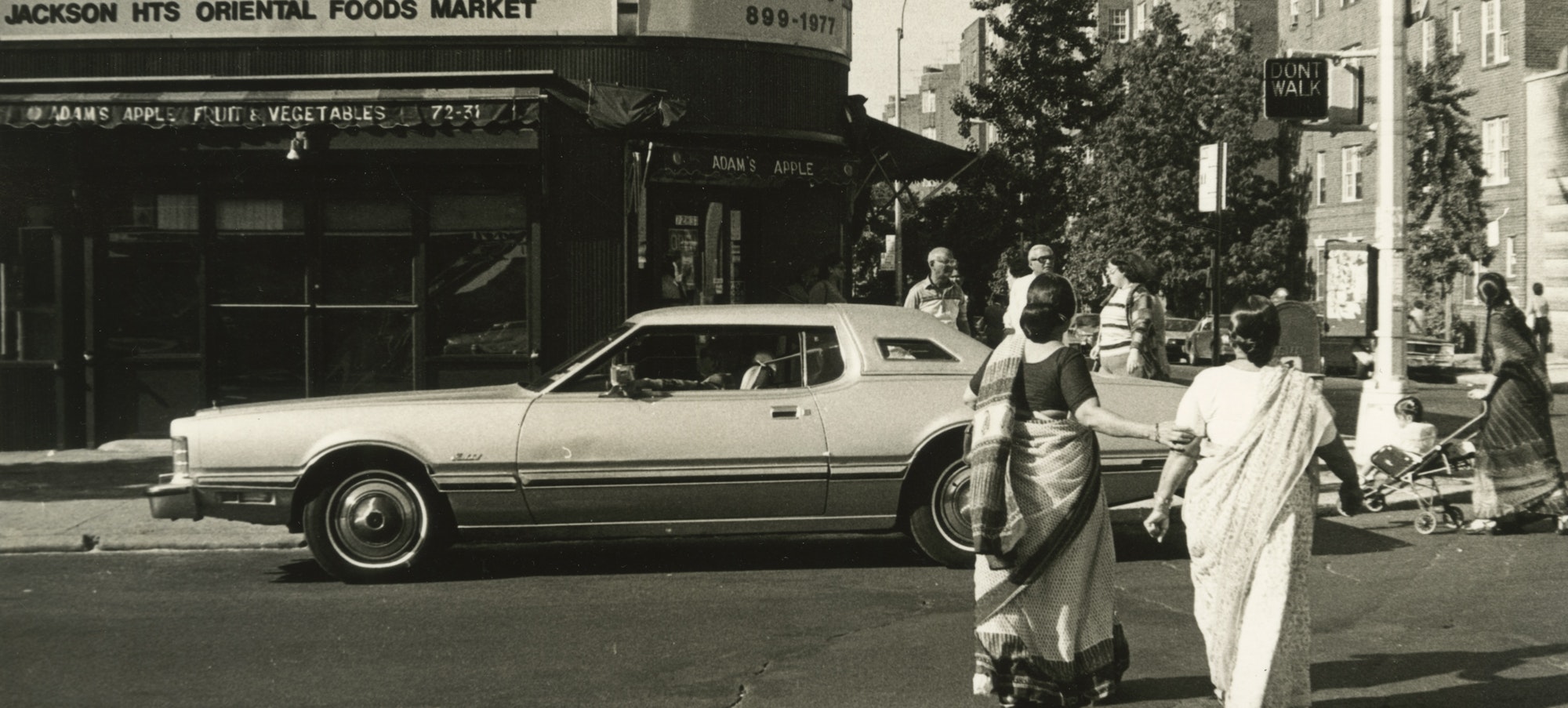 a black and white photo from 1984, showing a street in Jackson Heights queens, with a car passing by and two women in saris facing away from the camera.