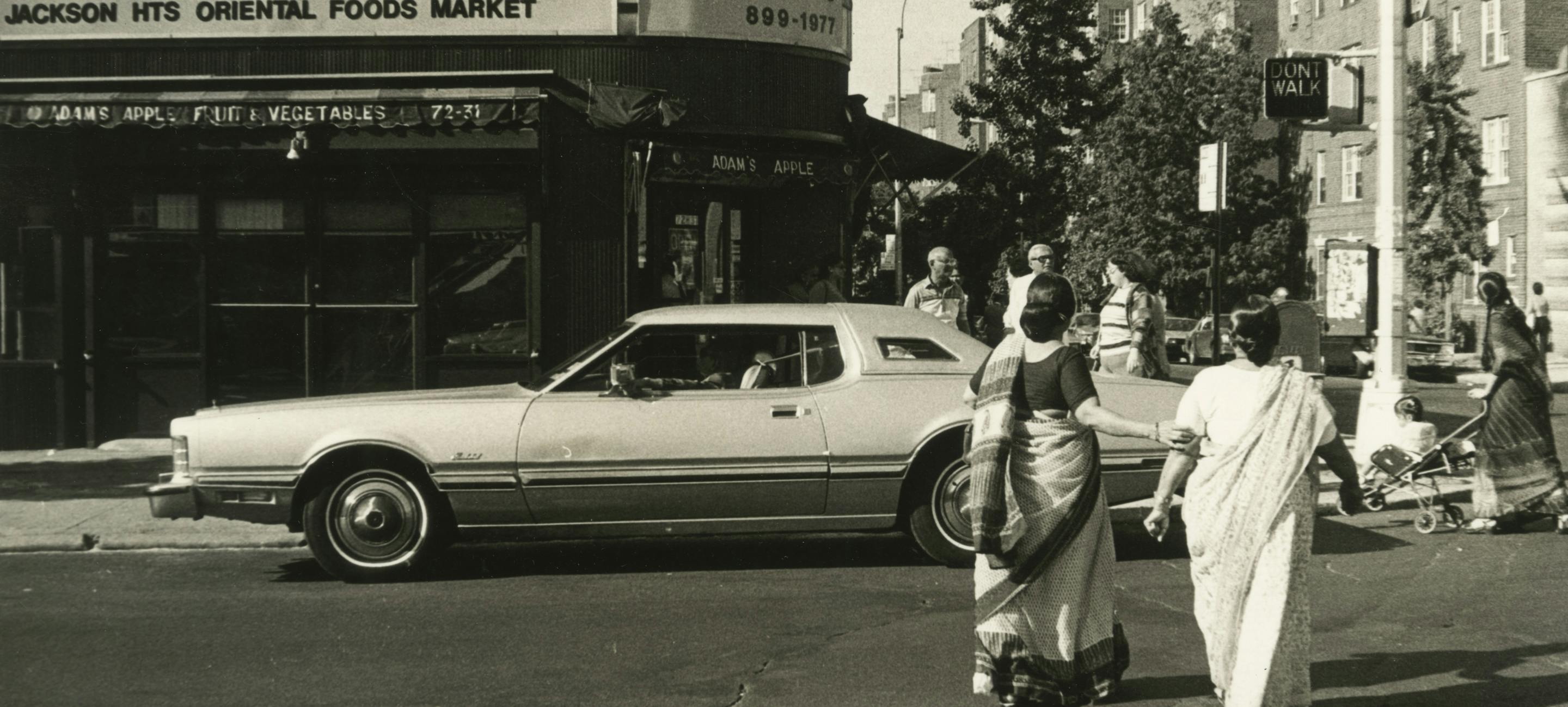 a black and white photo from 1984, showing a street in Jackson Heights queens, with a car passing by and two women in saris facing away from the camera.