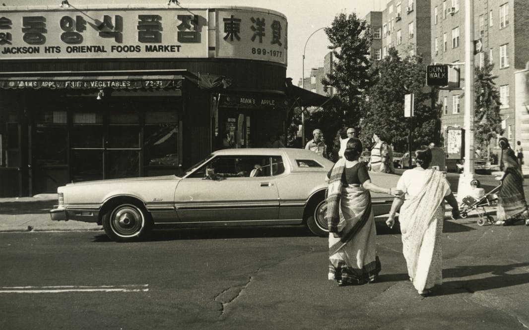 a black and white photo from 1984, showing a street in Jackson Heights queens, with a car passing by and two women in saris facing away from the camera.