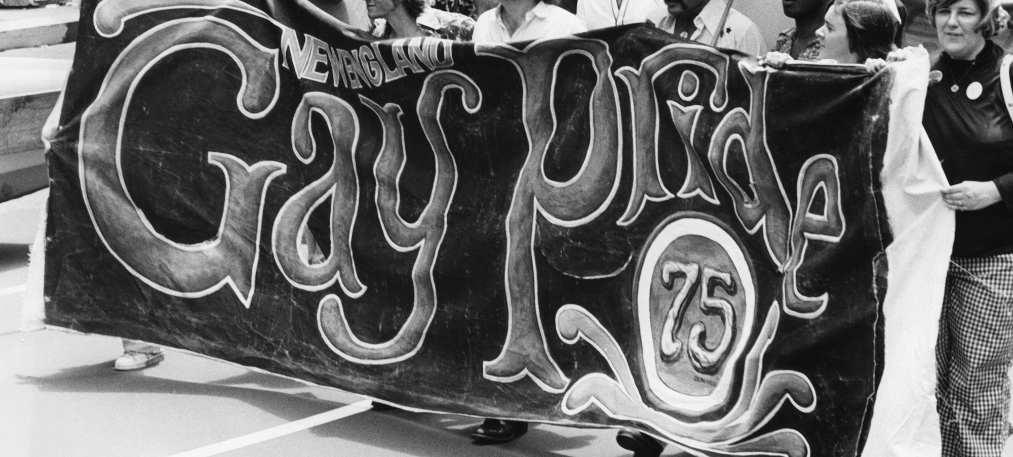 A line of participants at the 1975 Pride March in New York City carry a large banner reading "New England Gay Pride 75"