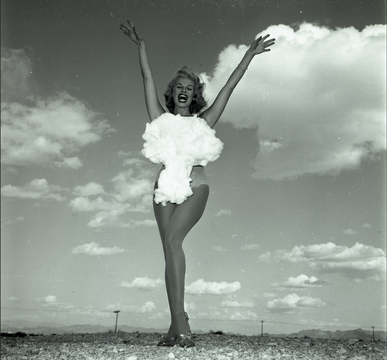 a black and white image of a woman in a bikini with a nuclear cloud over it raising her arms and smiling against a clear sky. Don English, Lee Merlin: “Miss Atomic Bomb,” 1957. Las Vegas News Bureau. Courtesy of LVCVA Archive, Las Vegas.