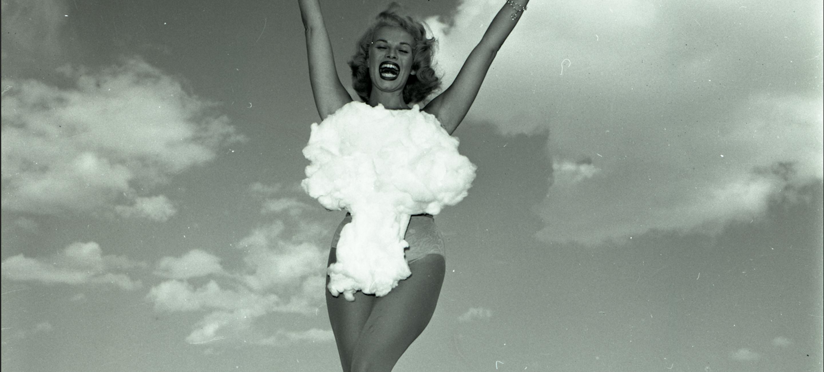 a black and white image of a woman in a bikini with a nuclear cloud over it raising her arms and smiling against a clear sky. Don English, Lee Merlin: “Miss Atomic Bomb,” 1957. Las Vegas News Bureau. Courtesy of LVCVA Archive, Las Vegas.