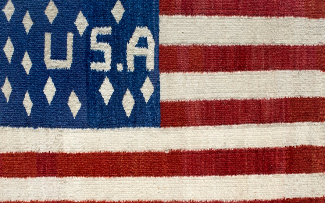 A woven American flag with a "USA" in the blue field and diamonds instead of stars. Diné Weaver, USA flag, late 19th century. Handspun wool, vegetal and native dyes. The New York Historical, Promised gift of Agnes Hsu-Tang and Oscar Tang, The Hsu-Tang Collection