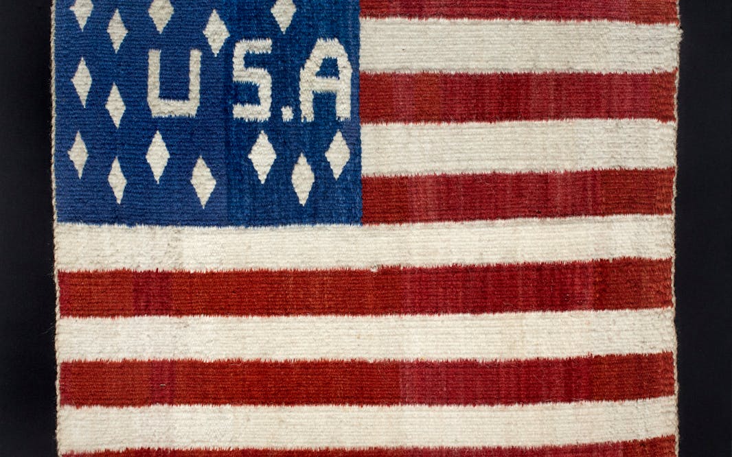 A woven American flag with a "USA" in the blue field and diamonds instead of stars. Diné Weaver, USA flag, late 19th century. Handspun wool, vegetal and native dyes. The New York Historical, Promised gift of Agnes Hsu-Tang and Oscar Tang, The Hsu-Tang Collection