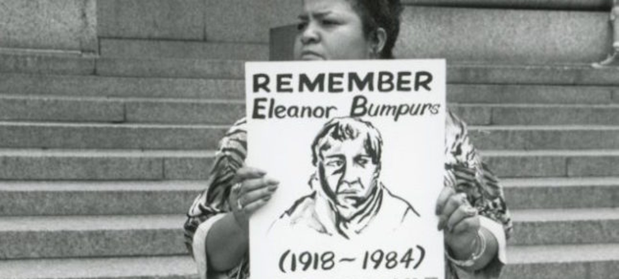 Woman in white dress standing on courthouse steps, holding an image of Eleanor Bumpurs