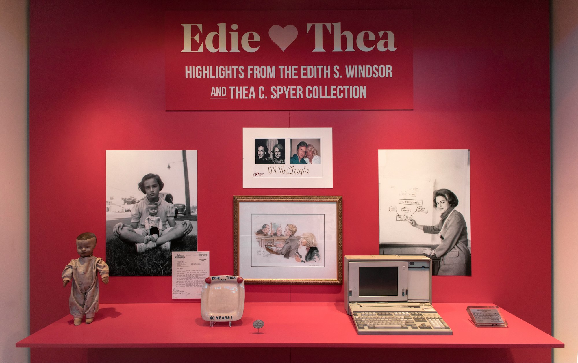 installation image of the Edie hearts Thea niche in objects tell stories, highlighting objects from the Edith S. Windsor and Thea C. Spyer Collection, like an older computer, a doll, a commemorative plate, and several photographs