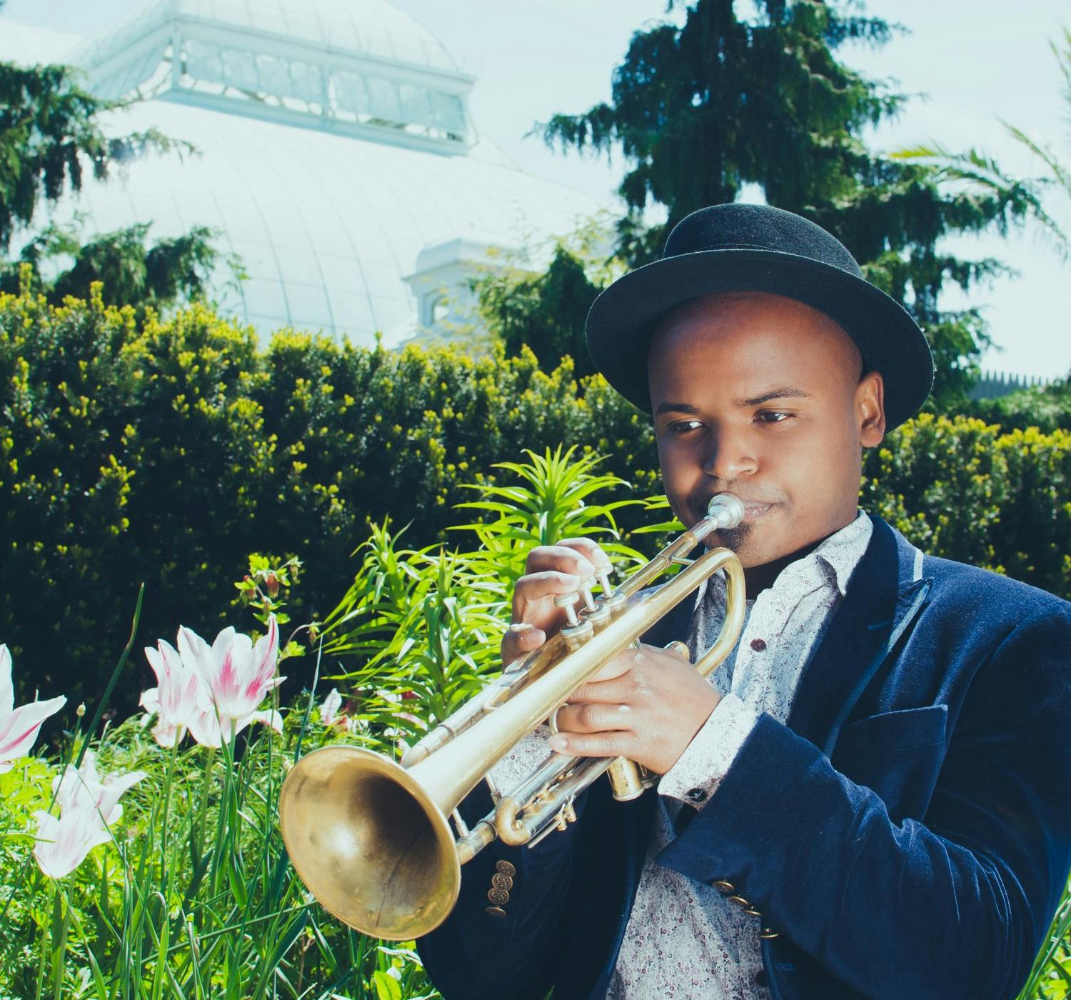the trumpet player alphonso Horne standing in a garden with his trumpet held to his lips.