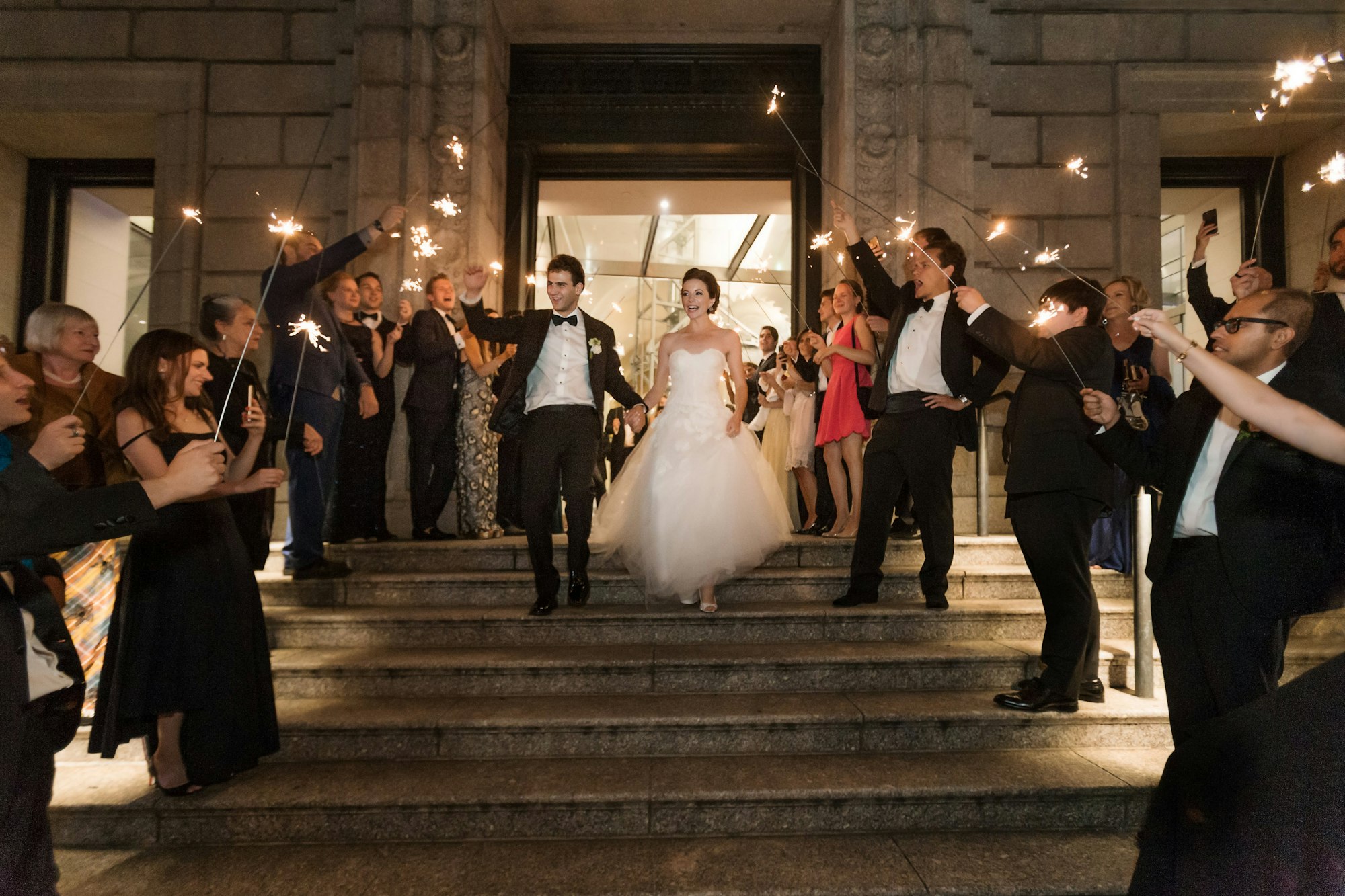 a newly married couple walk down the stairs of the New-York Historical Society while their guests cheer and hold sparklers