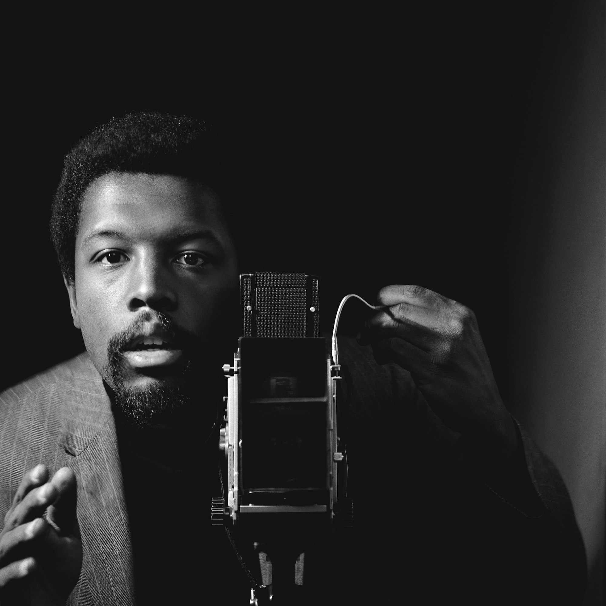 a black and white self-portrait of Kwame Brathwaite, a Black man with a goatee who's standing next to a camera.