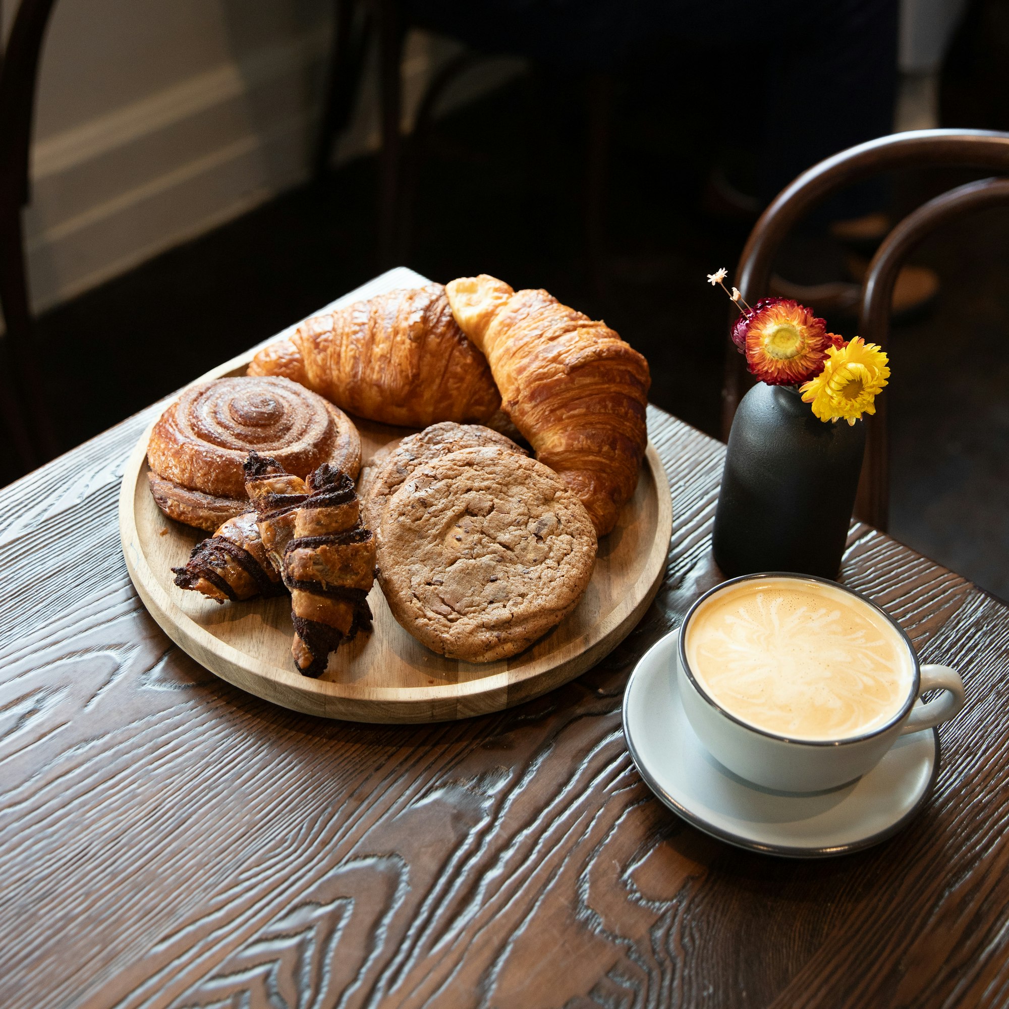 a plate of pastries on a table on Cafe 77, including cookies and croissants near a cappuccino in a white cup on a saucer and a vase of two small yellow flowers