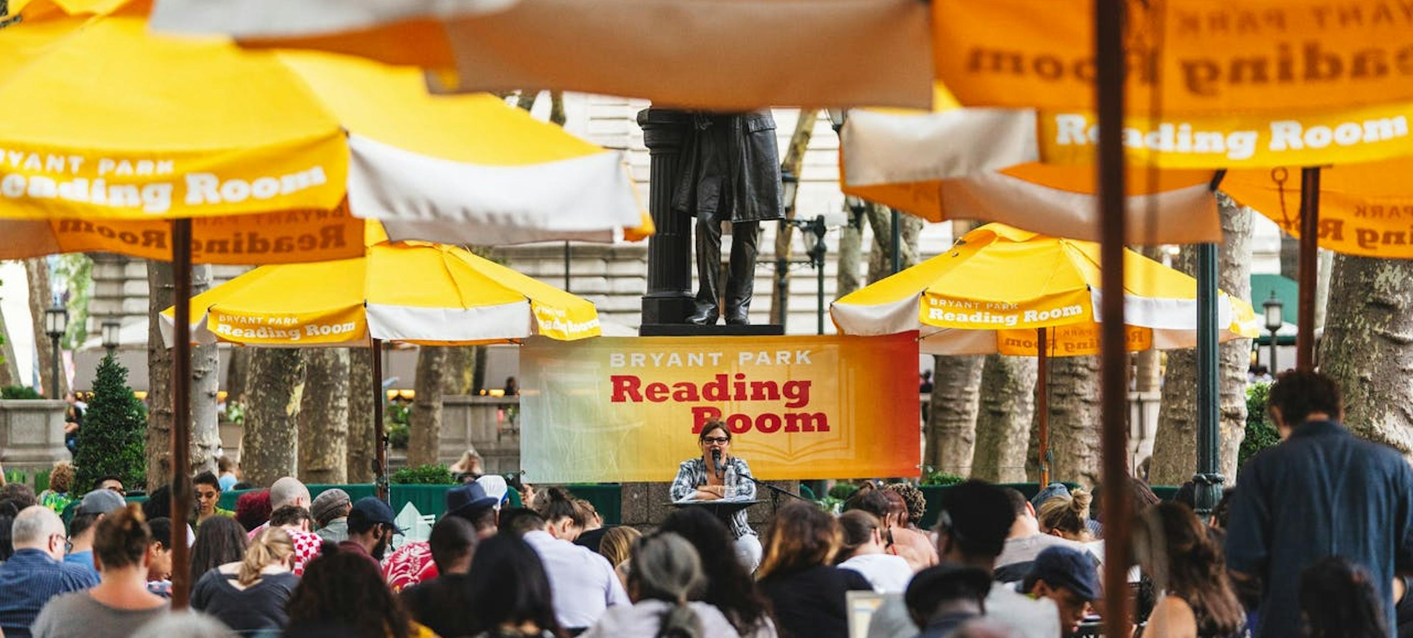 a speaker speaks in front of an audience at the bryant park reading room under yellow umbrellas